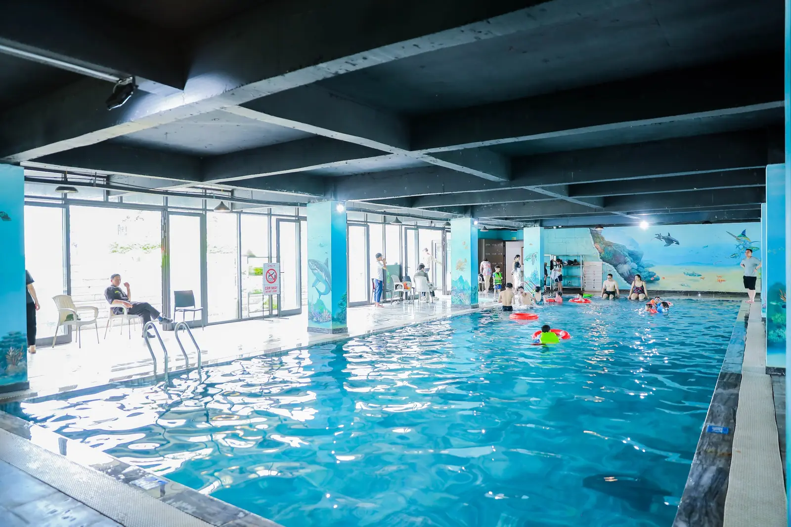 Bể bơi bốn mùa trong nhà ở Glory Resort: mặt nước xanh, cột và tường vẽ tranh đại dương, vài trẻ em bơi phao phía xa