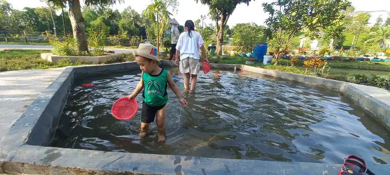 Bé trai cầm vợt đứng giữa bể bắt cá nước nông, phía sau là người lớn và vườn cây