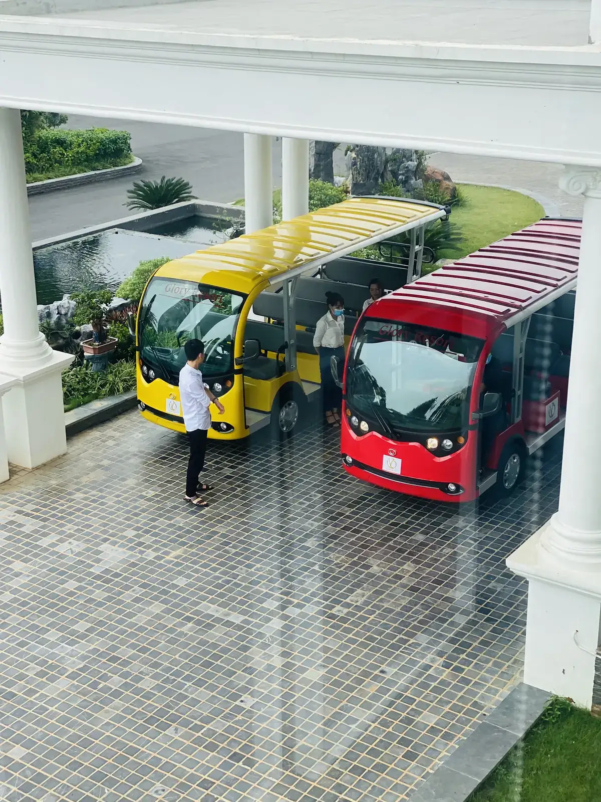 Hai xe điện nội khu màu vàng và đỏ chờ khách dưới sảnh có hàng cột trắng ở Glory Resort