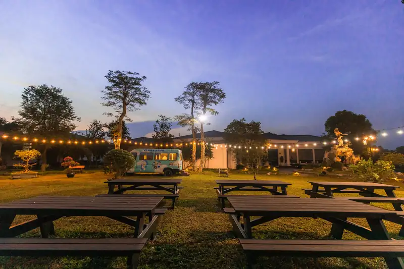 Khu BBQ ngoài trời Glory Resort lúc chạng vạng: bàn ghế gỗ dài trên bãi cỏ dưới dây đèn vàng