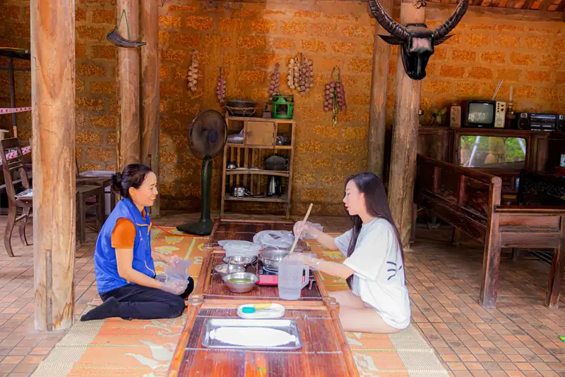 Hướng dẫn viên chỉ khách làm chè lam trên chiếu, trong gian nhà gỗ tường đá ong