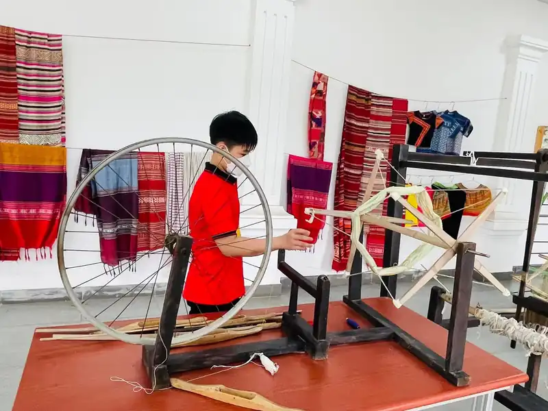 Bé trai thử quay sợi bên khung xe dệt thủ công, phía sau treo thổ cẩm nhiều màu