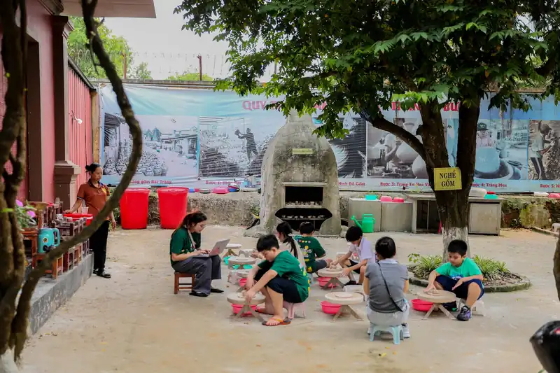 Trẻ em ngồi bàn xoay làm gốm dưới bóng cây, cạnh lò nung và bức ảnh làng gốm