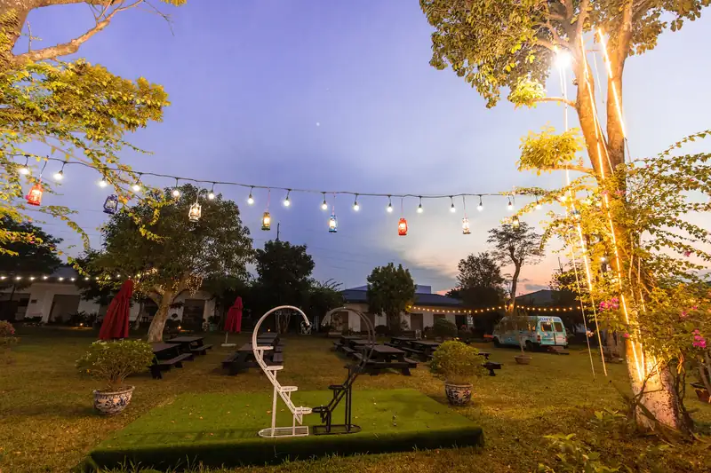 Khu BBQ ngoài trời lúc chạng vạng, dây đèn treo giữa tán cây và bàn gỗ trên bãi cỏ