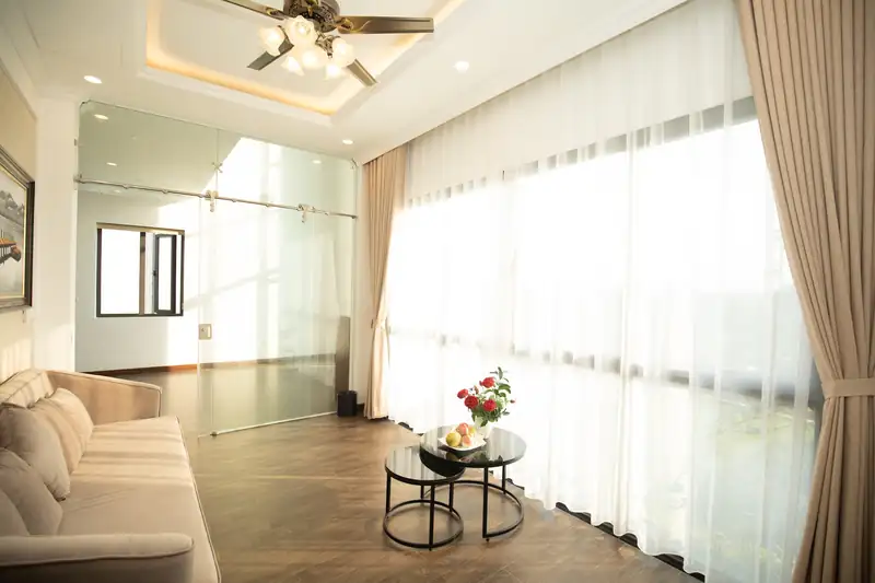 Phòng khách Osaka Villa với sofa nỉ, bàn trà và rèm voan trắng trải dài theo vách kính