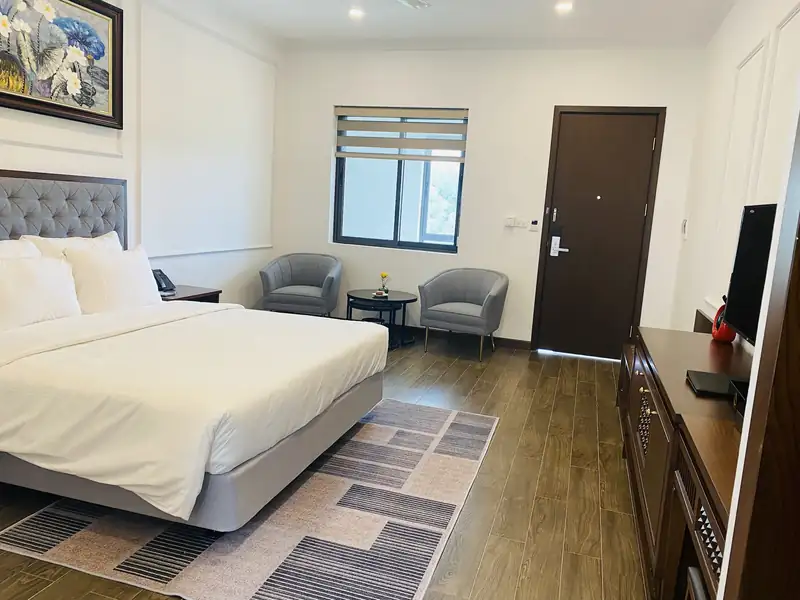 Phòng giường King 1,8 m nhìn về phía cửa: đôi ghế bành xám, bàn tròn, cửa sổ và tủ ti vi gỗ