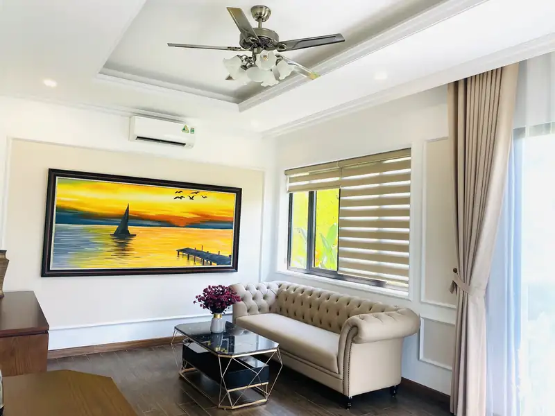 Phòng khách LakeSide Villa với sofa kem, bàn trà, tranh thuyền buồm và quạt trần