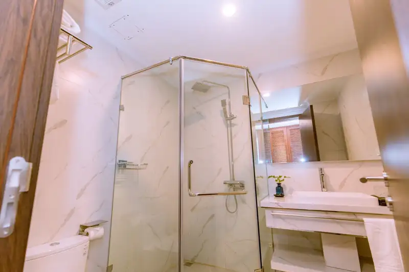 Phòng tắm tòa Pool View: vách kính vòi sen, tường đá vân trắng, lavabo và gương lớn