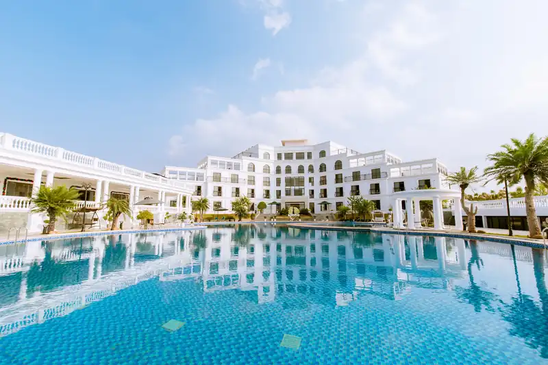 Tòa Pool View Glory Resort nhìn qua mặt bể bơi vô cực xanh, hàng cọ và trời nắng