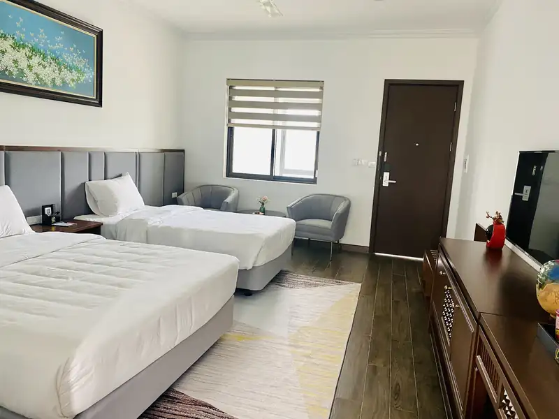Phòng hai giường lớn nhỏ nhìn về phía cửa: đôi ghế bành xám, cửa sổ rèm cuốn và tủ gỗ