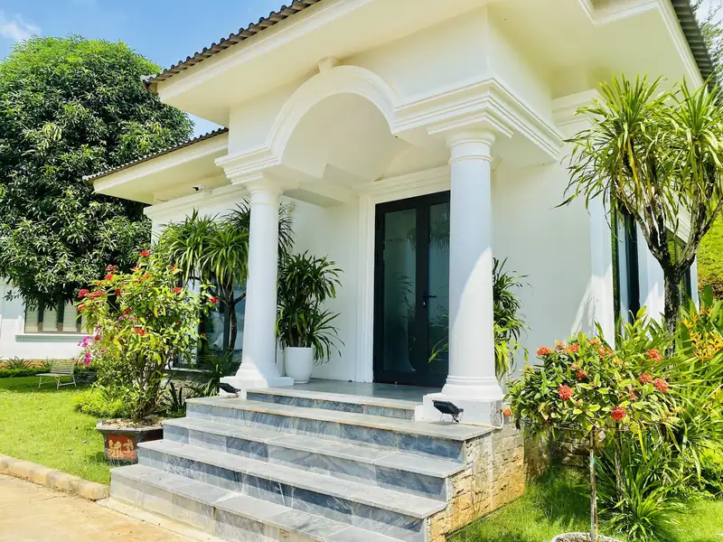Hiên trước biệt thự với cột tròn, bậc đá xám và khóm hoa đỏ hai bên lối vào